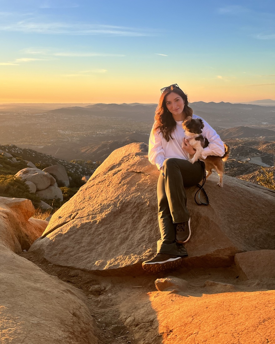 Cristina Panzarini sitting on a boulder at sunset, holding her dog Saki, with hills and a valley behind them